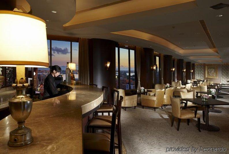 Ambassador Hotel Гаосюн Ресторан фото The photo depicts an elegant bar or lounge area within a high-rise setting. The interior features warm lighting, with a large lamp illuminating the bar counter. There is a bartender seated at the bar, and the space includes plush chairs and tables ar