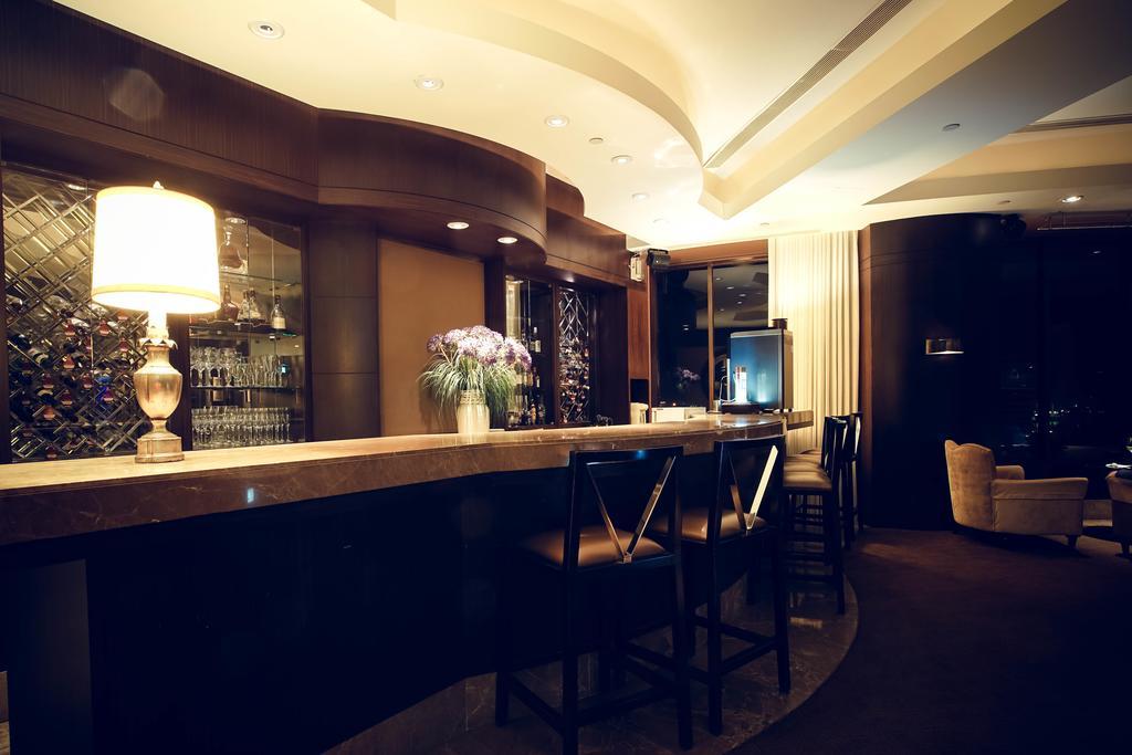 Ambassador Hotel Гаосюн Экстерьер фото The photo shows a modern and stylish bar area. The bar is made of a smooth stone surface and features a backlit shelf displaying various bottles of liquor. There is a lamp on the bar providing warm lighting, and a vase with flowers adds a decorative 