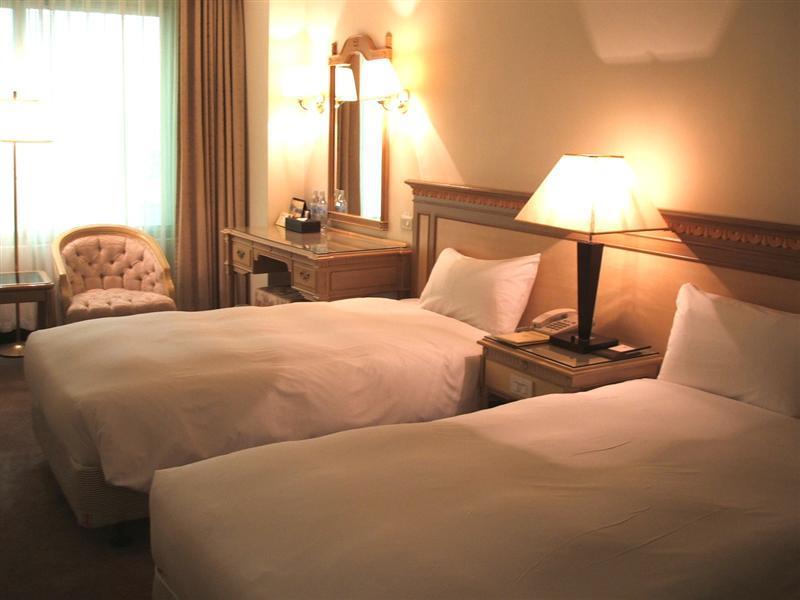 Ambassador Hotel Гаосюн Экстерьер фото The photo shows a cozy hotel room with two single beds, each covered with white bedding. There is a lamp on a nightstand between the beds. A wooden dresser with a mirror is against the wall, and a comfortable armchair is placed nearby. Soft lighting 
