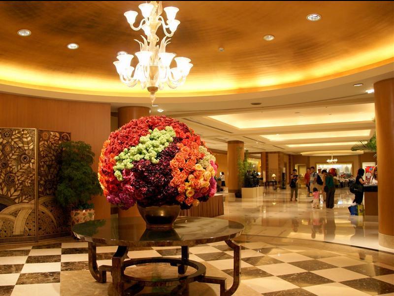 Ambassador Hotel Гаосюн Экстерьер фото The photo shows the interior of a spacious lobby area, characterized by elegant design elements. In the center, there is a large round floral arrangement made up of vibrant flowers in various colors, displayed in a decorative vase on a dark, round ta