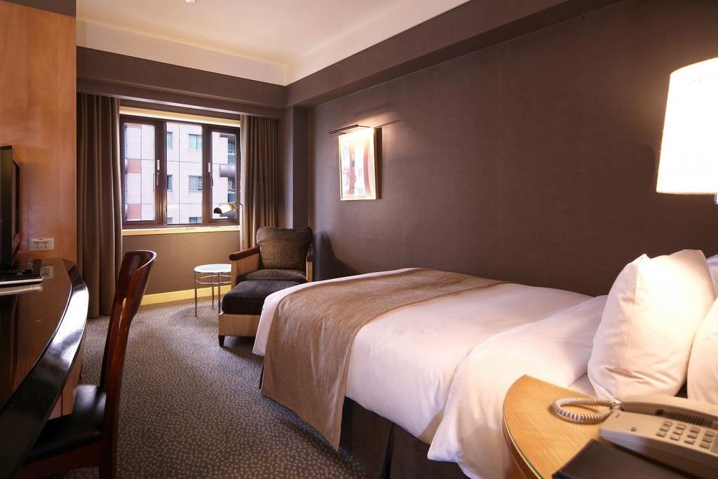 Ambassador Hotel Гаосюн Экстерьер фото The photo shows a hotel room featuring a neatly made single bed with a light-colored blanket. There is a comfortable chair in the corner alongside a small round table. A large window allows natural light to enter the room, and the walls are painted i