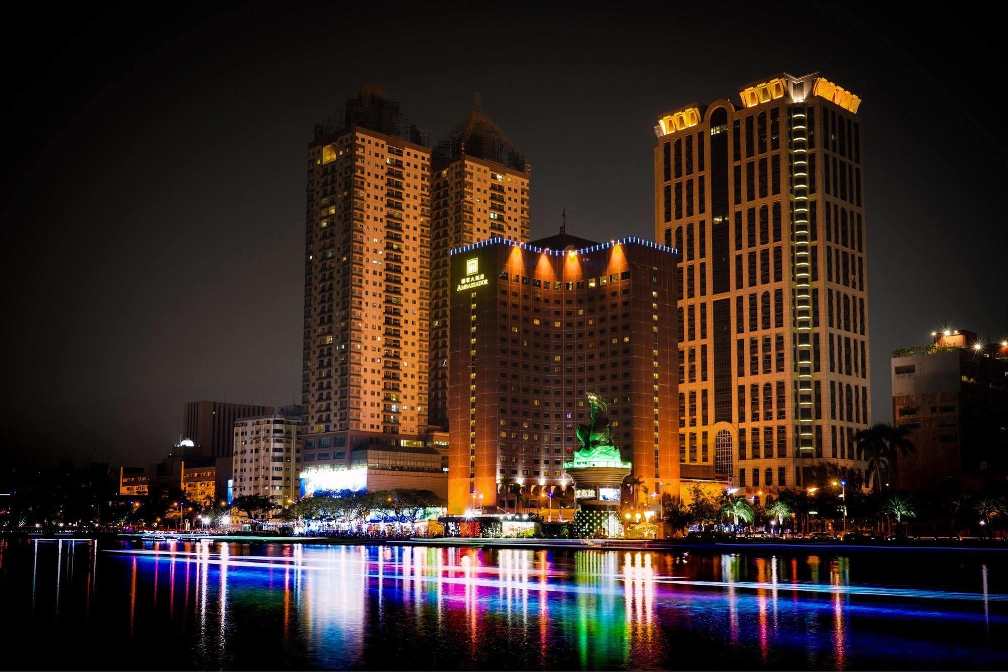 Ambassador Hotel Гаосюн Экстерьер фото The photo depicts a vibrant nighttime cityscape featuring several tall buildings illuminated by colorful lights. Prominent structures include a large hotel and other high-rise buildings with illuminated facades. In front of these buildings, there app