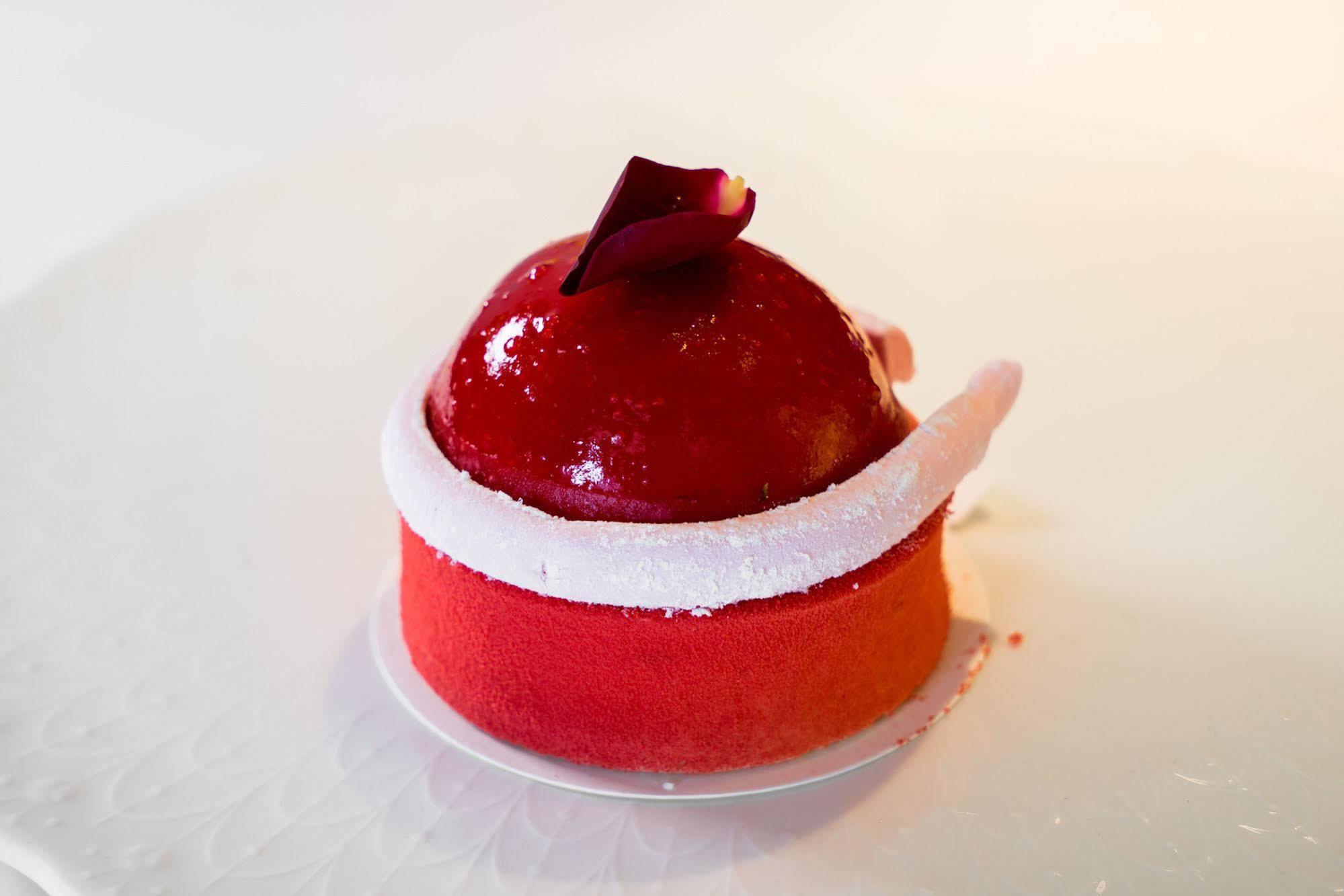 Ambassador Hotel Гаосюн Экстерьер фото The photo shows a beautifully crafted dessert that appears to be a mousse or cake. It has a glossy red coating, shaped like a dome on top, with a small decorative rose petal placed on it. The base of the dessert features a gradient of colors, transit