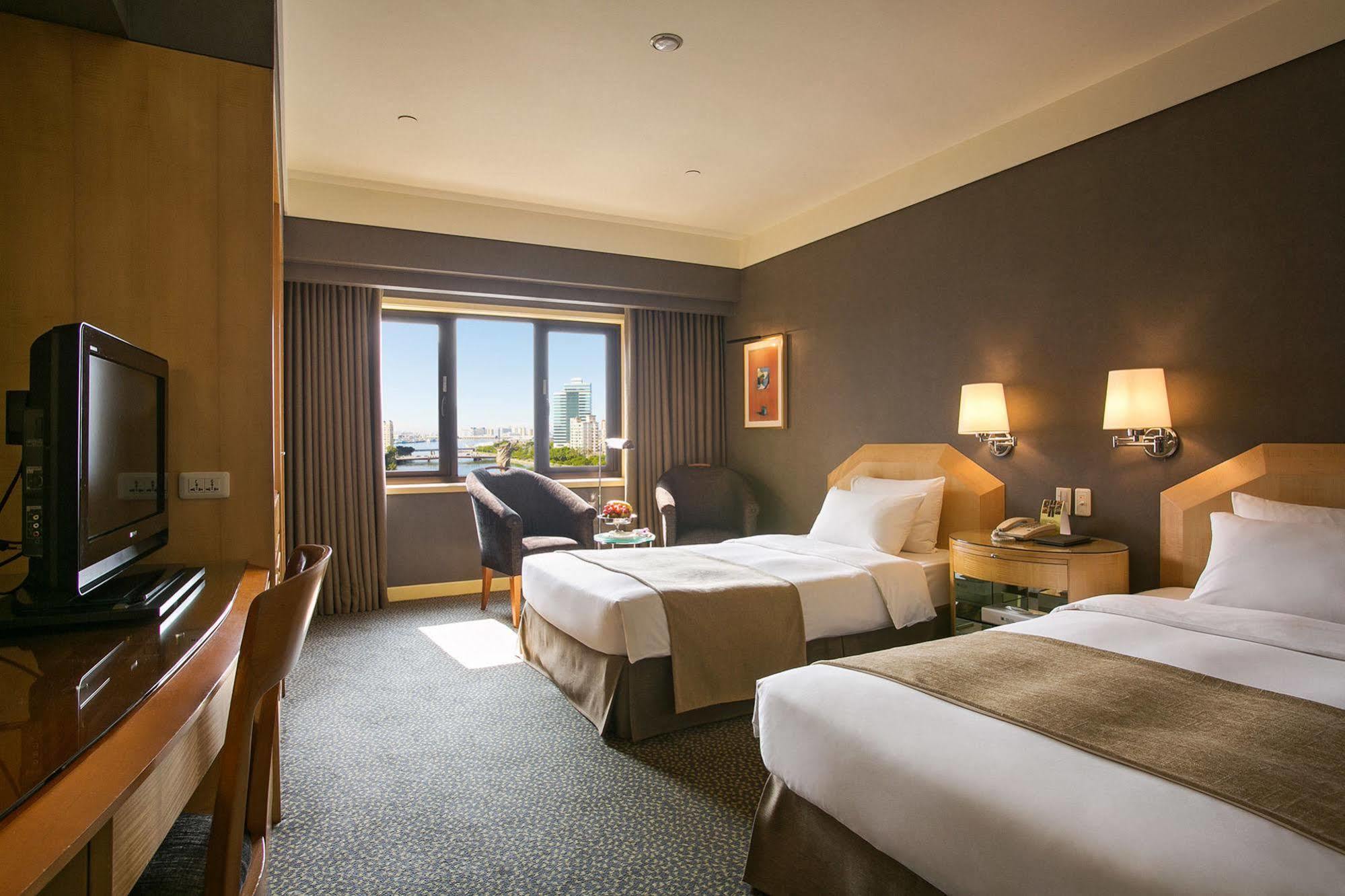 Ambassador Hotel Гаосюн Экстерьер фото The photo shows a hotel room with two beds, one of which is a double bed and the other appears to be a single. The room has a modern design with a neutral color palette, featuring wooden furniture, a TV unit, and a small desk and chair. There are two