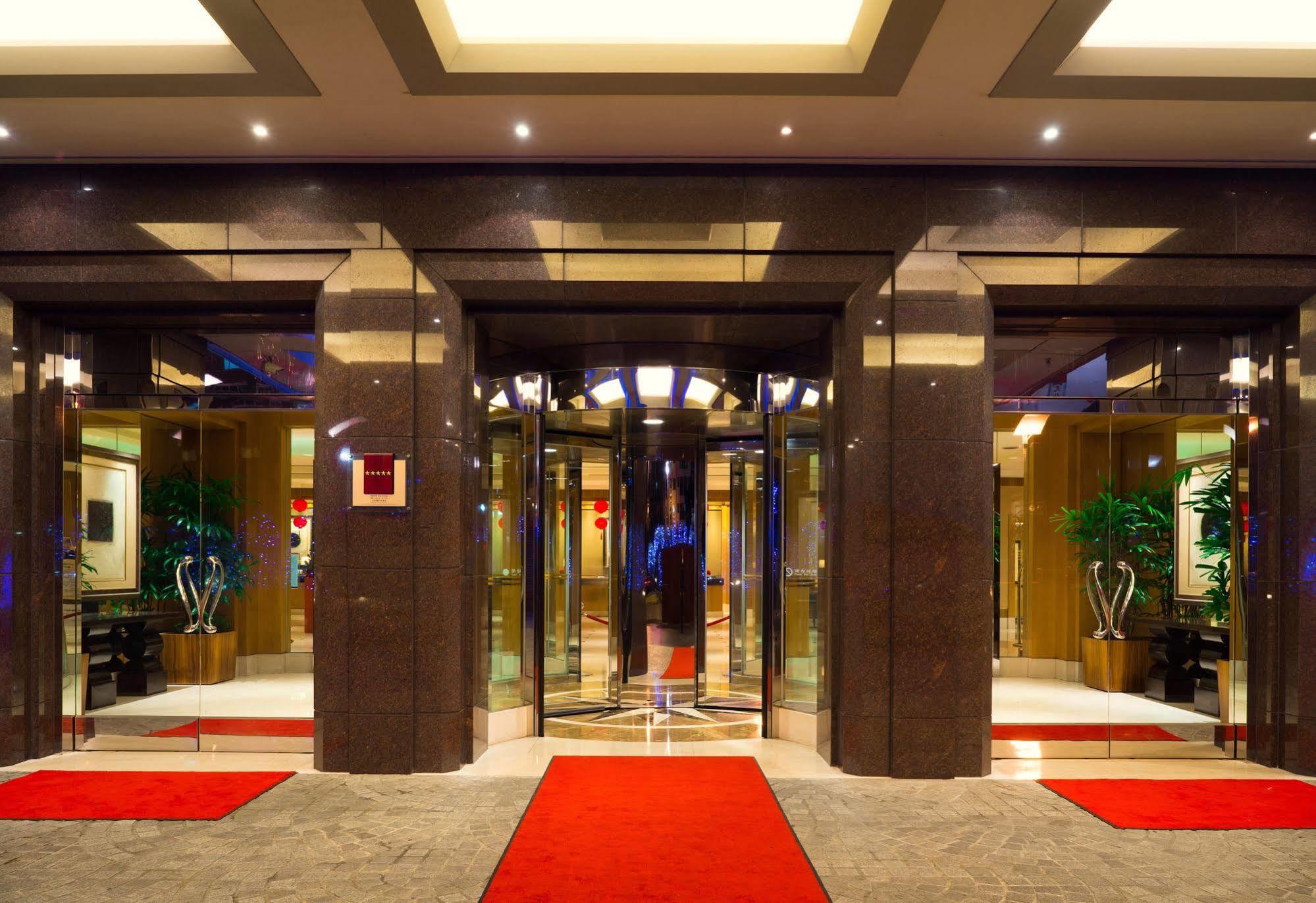 Ambassador Hotel Гаосюн Экстерьер фото The photo shows the entrance of a modern hotel or building. It features large glass doors in a sleek frame, creating an inviting entrance. The floor is covered with a plush red carpet leading up to the doors. Inside, there are decorative plants on ei