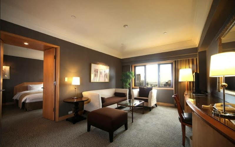 Ambassador Hotel Гаосюн Экстерьер фото The photo shows a stylish hotel room with modern décor. There is a comfortable seating area featuring a white sofa and brown ottoman, along with a glass coffee table. A side table with a lamp is positioned next to the sofa. The room has a large windo