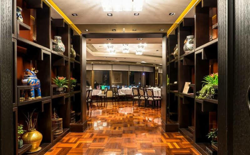 Ambassador Hotel Гаосюн Экстерьер фото The photo shows an elegant restaurant interior. In the foreground, there are dark wooden shelves filled with decorative items, such as vases and small sculptures, along with plants. The floor is made of polished wood, featuring a geometric pattern. I